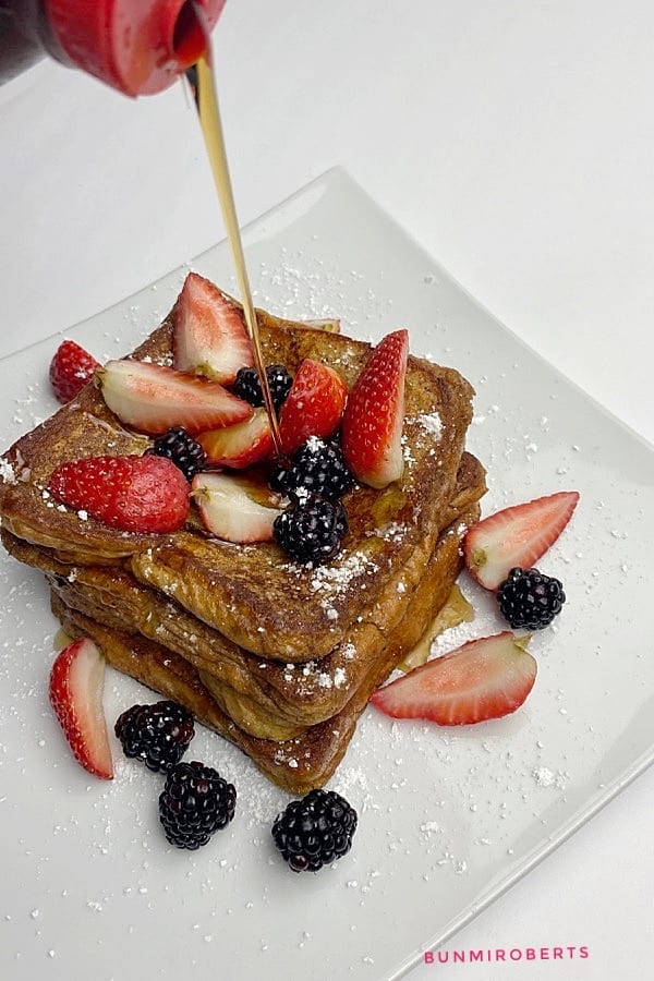 A picture of maple syrup being poured over a stack of cinnamon French toast served with strawberries, blackberries and powdered sugar